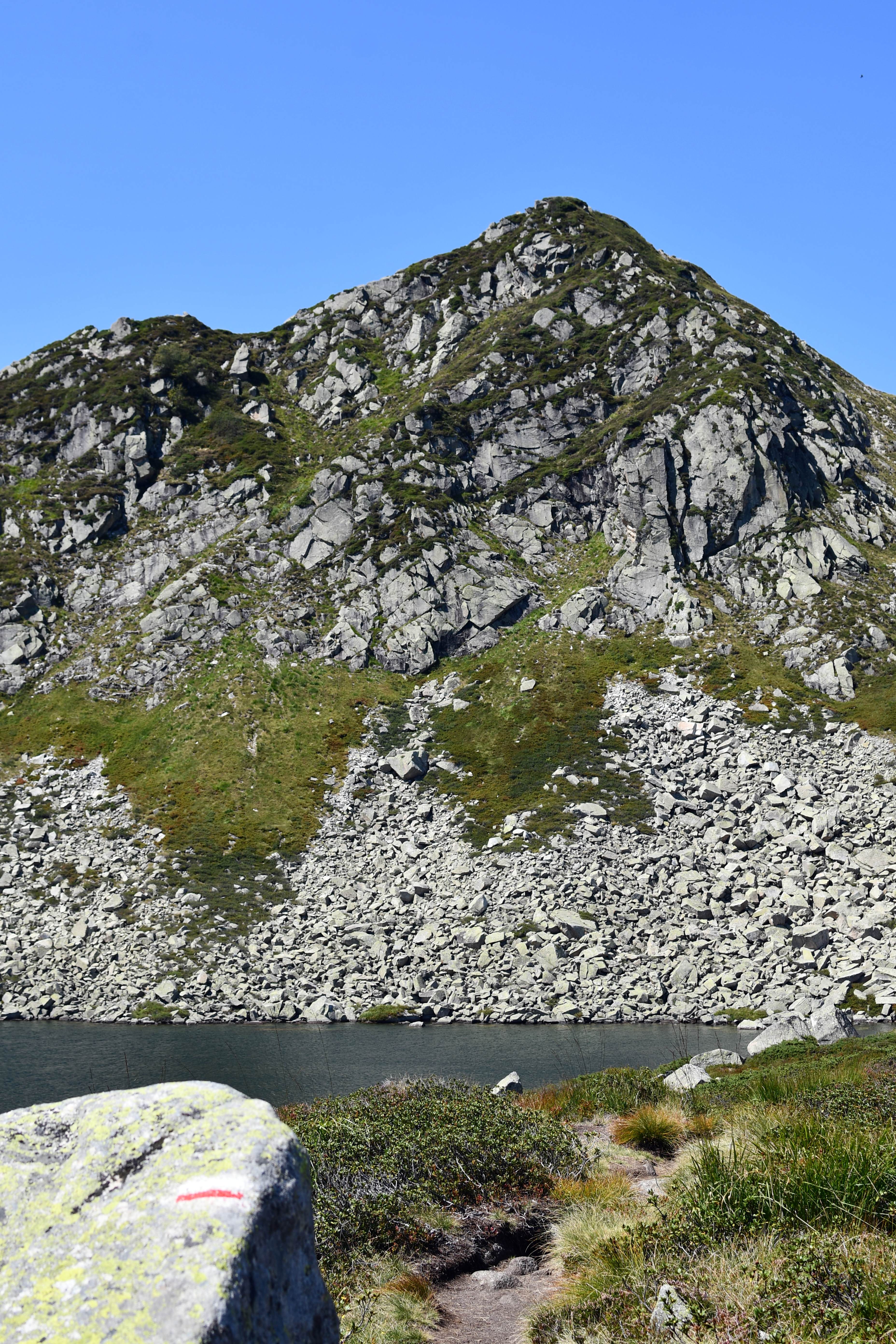 Un étang de montagne avec un sentier devant et une montagne caillouteuse derrière